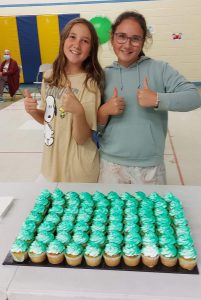Two female students give the thumbs up in front of a tray of cupcakes with green icing